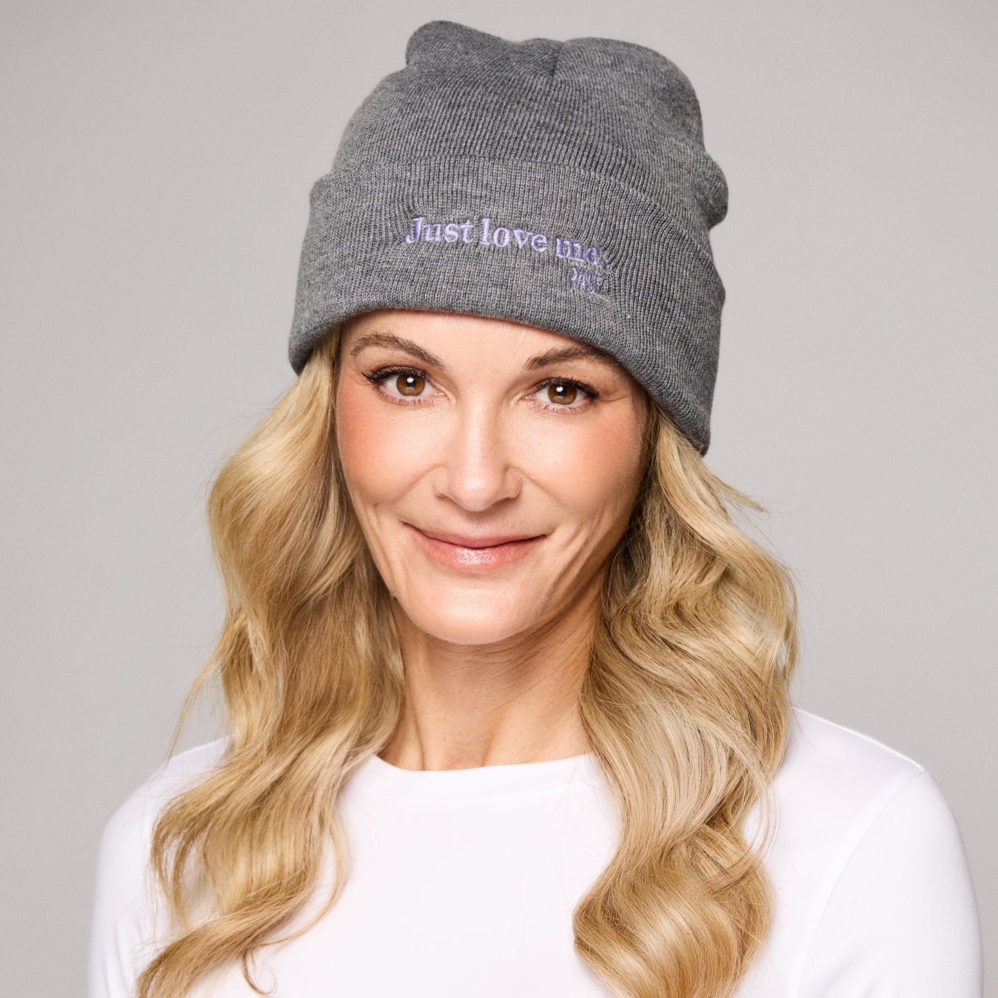 Woman wearing a beanie hat in color grey. The hat has a 'Just Love Me' message in purple.